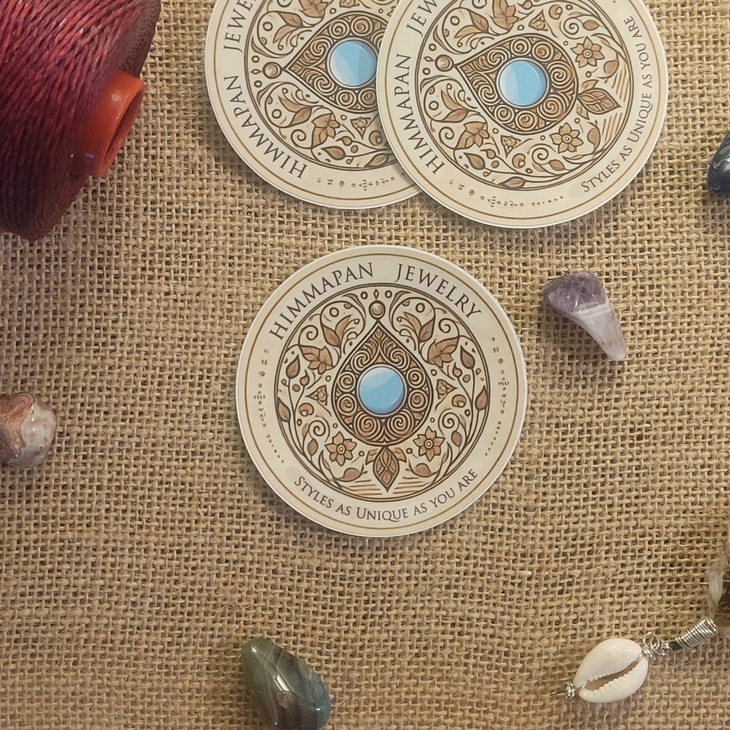 Himmapan Jewelry Logo Sticker displayed on a burlap surface with decorative stones.