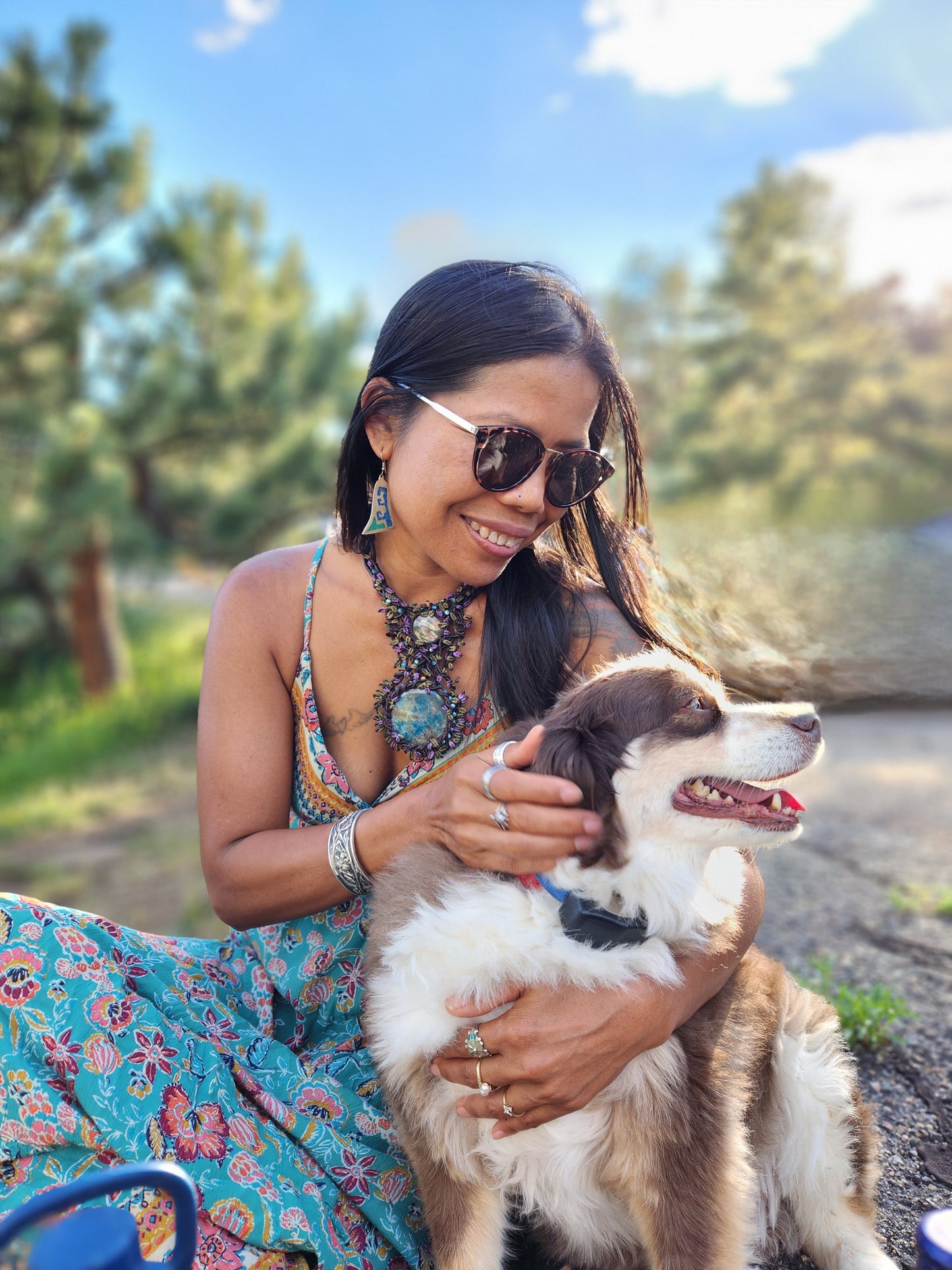Woman in a colorful dress sitting outdoors with a brown and white dog, smiling while wearing sunglasses and jewelry.
