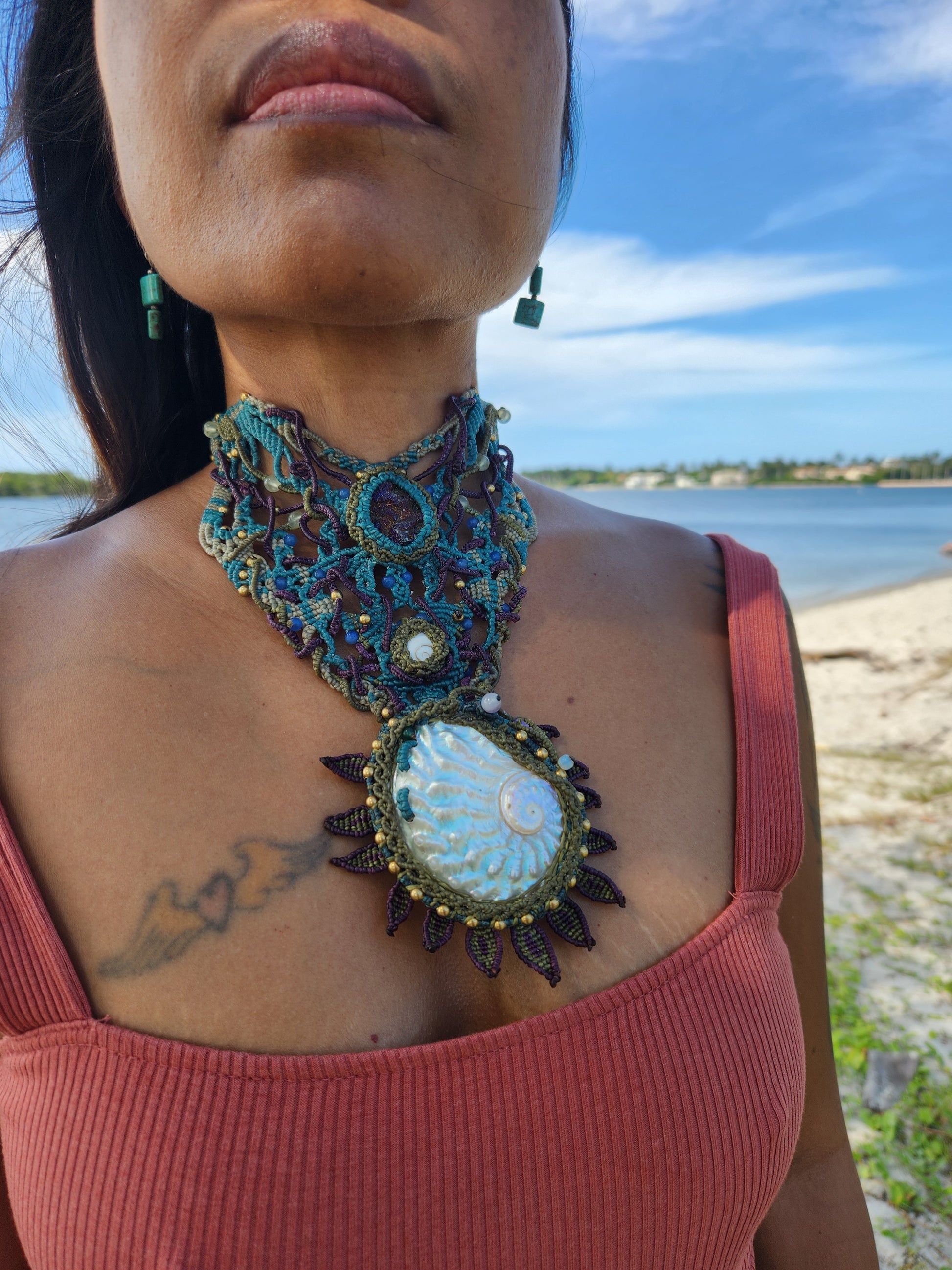 Shell Pendant worn as an ornate choker featuring a large abalone centerpiece and multicolored cord, set against a beach backdrop.