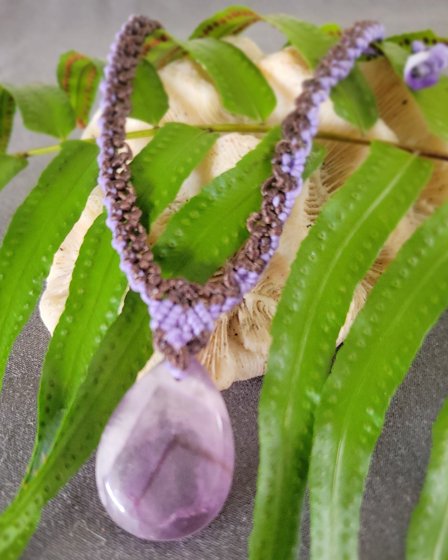 Amethyst Pendant – 'Purple Rain' Teardrop Crystal Pendant Necklace on macramé chain surrounded by green leaves.