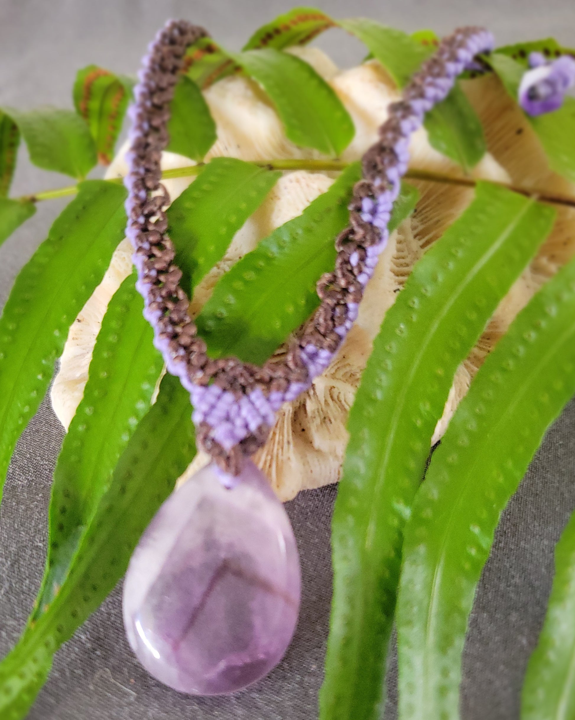 Amethyst Pendant – 'Purple Rain' Teardrop Crystal Pendant Necklace on macramé chain surrounded by green leaves.